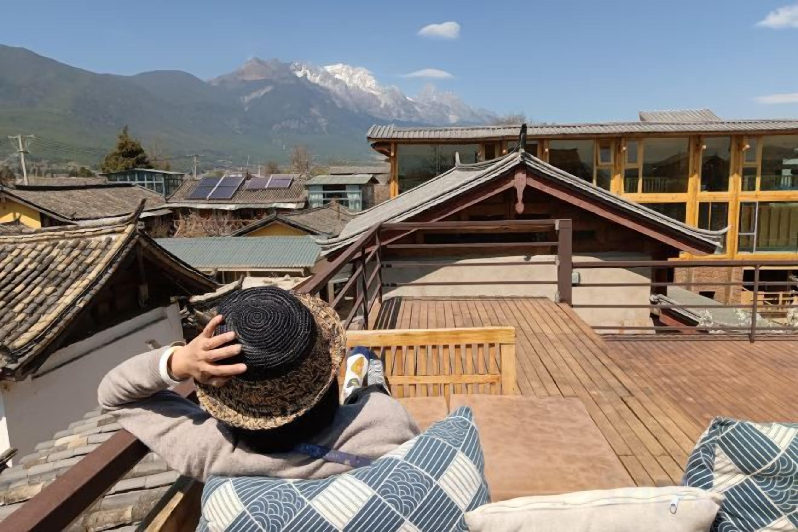Una persona descansando en una terraza de madera con vista a edificios tradicionales y una montaña al fondo.