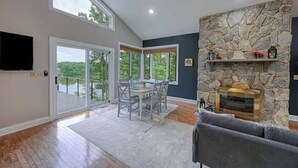 A spacious living area with a stone fireplace, a dining table with chairs, and large windows offering a view of the outdoors. - Stepping Stone Cove :: Lakefront w/ Dock, Fireplaces & Hot Tub (Mineral)