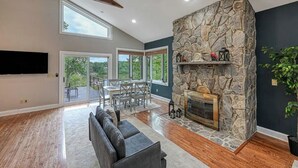 A spacious living room with a stone fireplace, a ceiling fan, and a large window overlooking a deck. - Stepping Stone Cove :: Lakefront w/ Dock, Fireplaces & Hot Tub (Mineral)