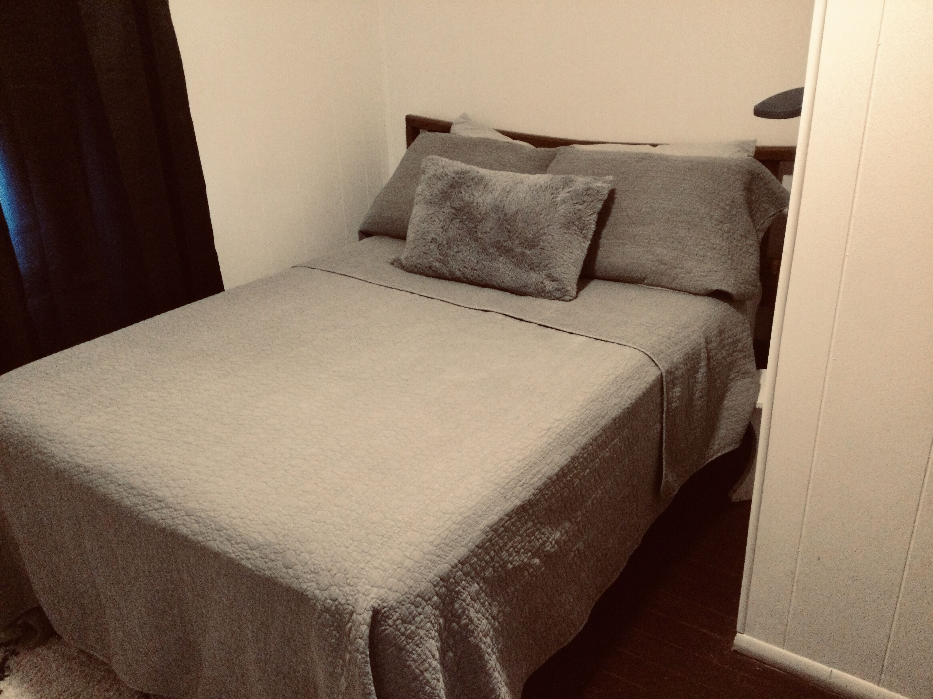 Una cama pequeña y bien hecha con una colcha y almohadas grises, ubicada en una esquina de una habitación con paredes blancas y suelo oscuro.
