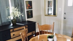 Un comedor con una mesa de madera redonda rodeada por seis sillas de madera. A la izquierda, un aparador oscuro con cajones. La habitación tiene una alfombra estampada y un espejo grande en la pared sobre la puerta.