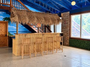 A bar area with a thatched roof, bamboo furniture, and a view of a staircase and greenery.