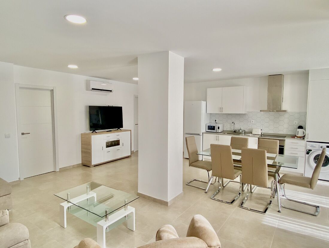 Una cocina moderna con gabinetes blancos, un comedor con mesa de vidrio y una sala de estar con sofá y mesa de centro.