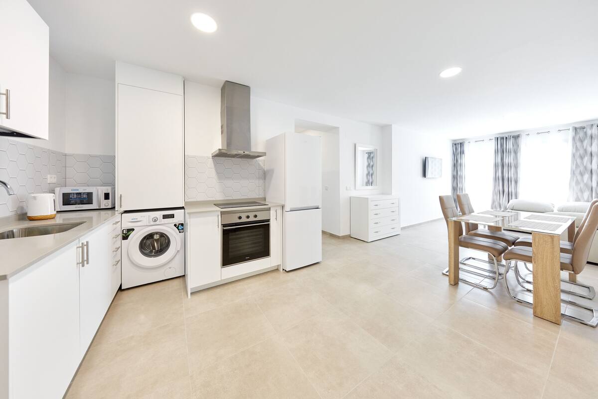 Een moderne keuken met een ingebouwde wasmachine, oven en kookplaat, en een eethoek met een tafel en stoelen.