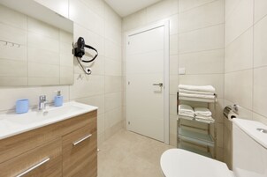 Un baño moderno con lavamanos blanco, tocador de madera, un secador de pelo, inodoro y un perchero para toallas.