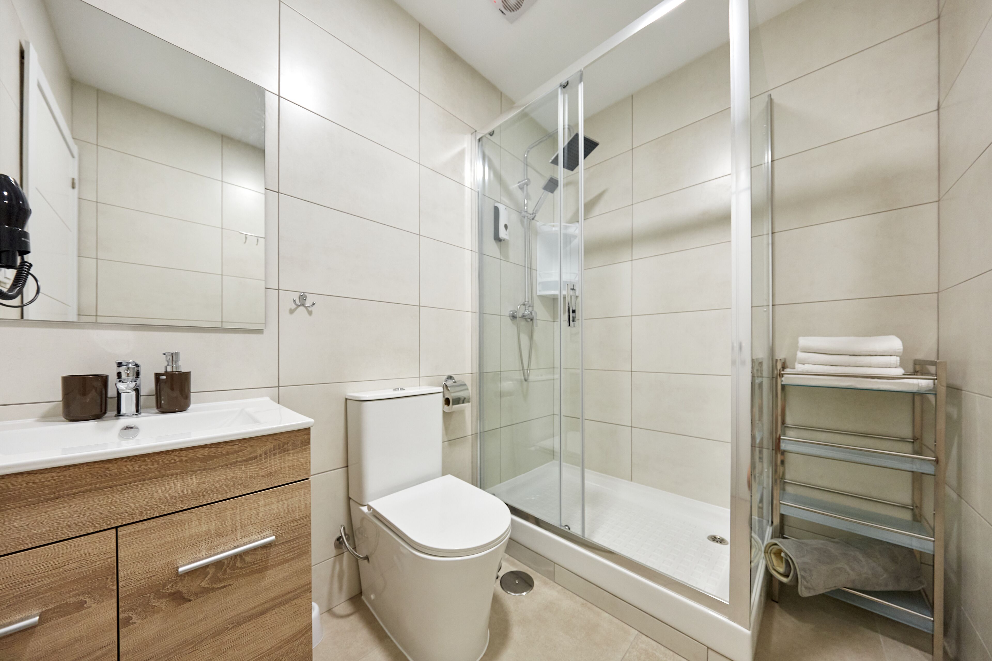 Un baño moderno con ducha de vidrio, inodoro blanco, tocador de madera con cajones y repisa de vidrio con toallas.