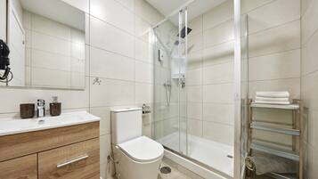 Un baño moderno con ducha de vidrio, inodoro blanco, tocador de madera con cajones y repisa de vidrio con toallas.
