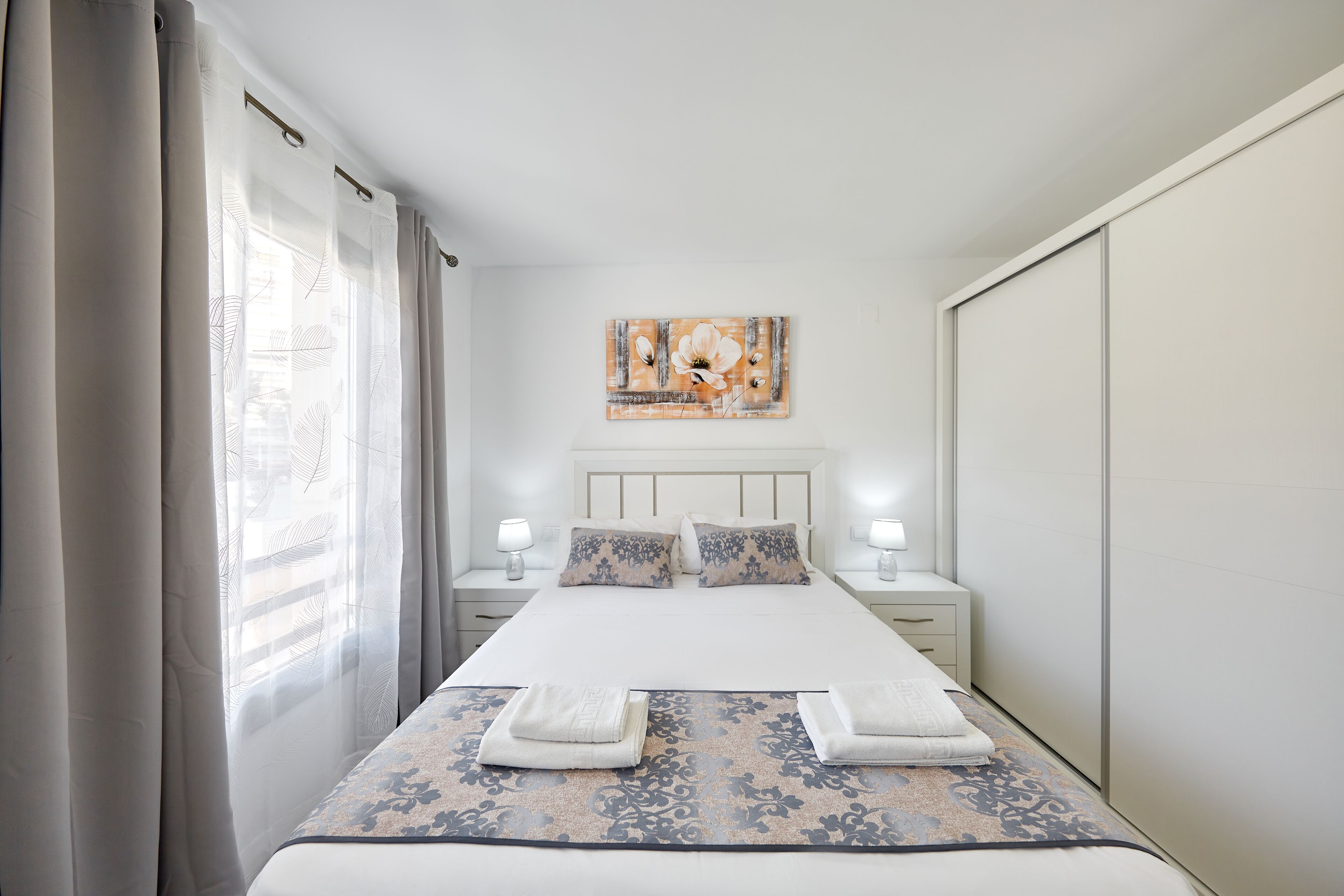 Una habitación de hotel ordenada con una cama grande, dos mesitas de noche con lámparas, un cabecero con una obra de arte floral y un armario.