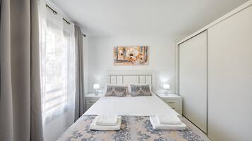 Una habitación de hotel ordenada con una cama grande, dos mesitas de noche con lámparas, un cabecero con una obra de arte floral y un armario.