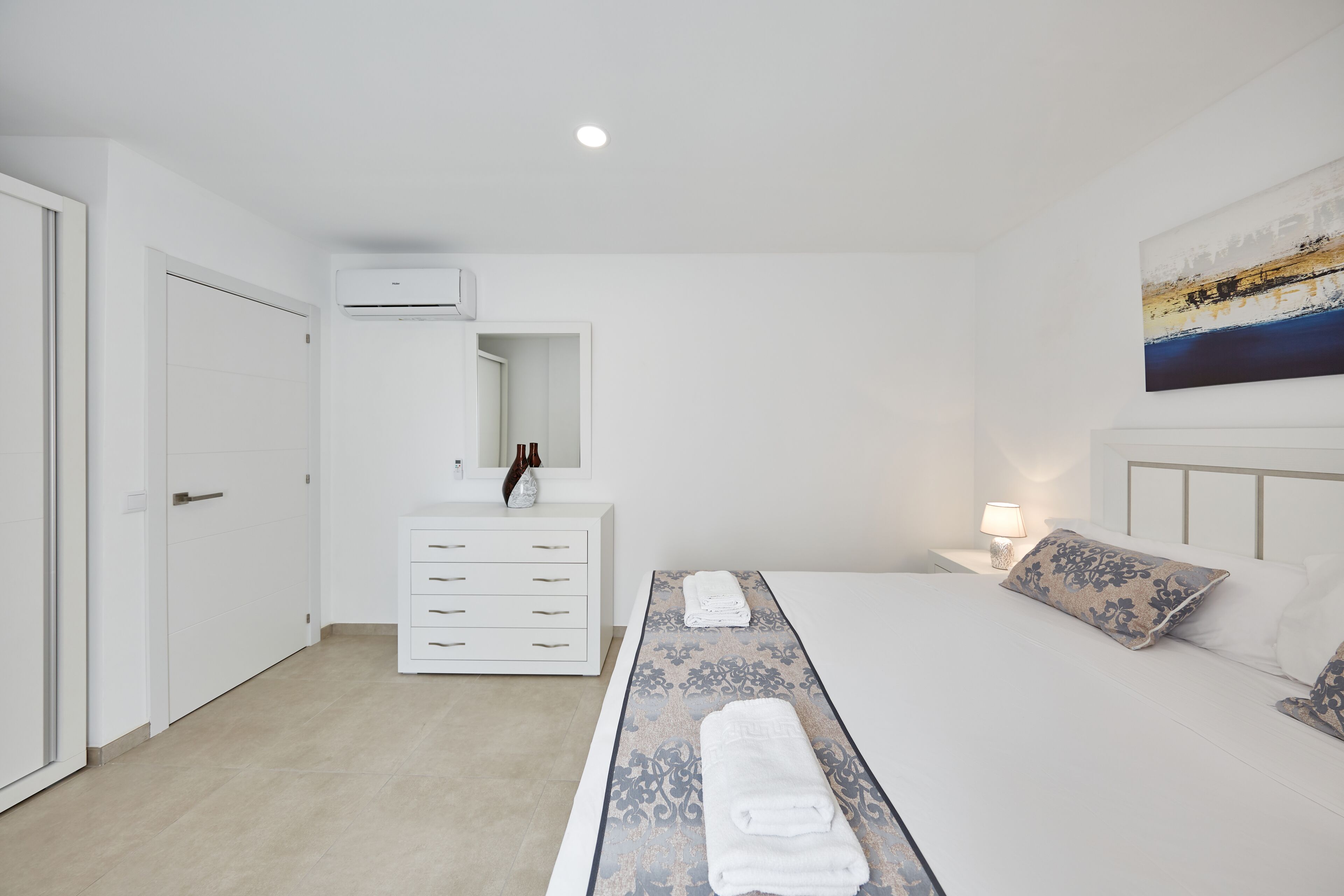 A modern hotel room with a neatly made bed, a bedside table with a lamp, a dresser, and a large window.