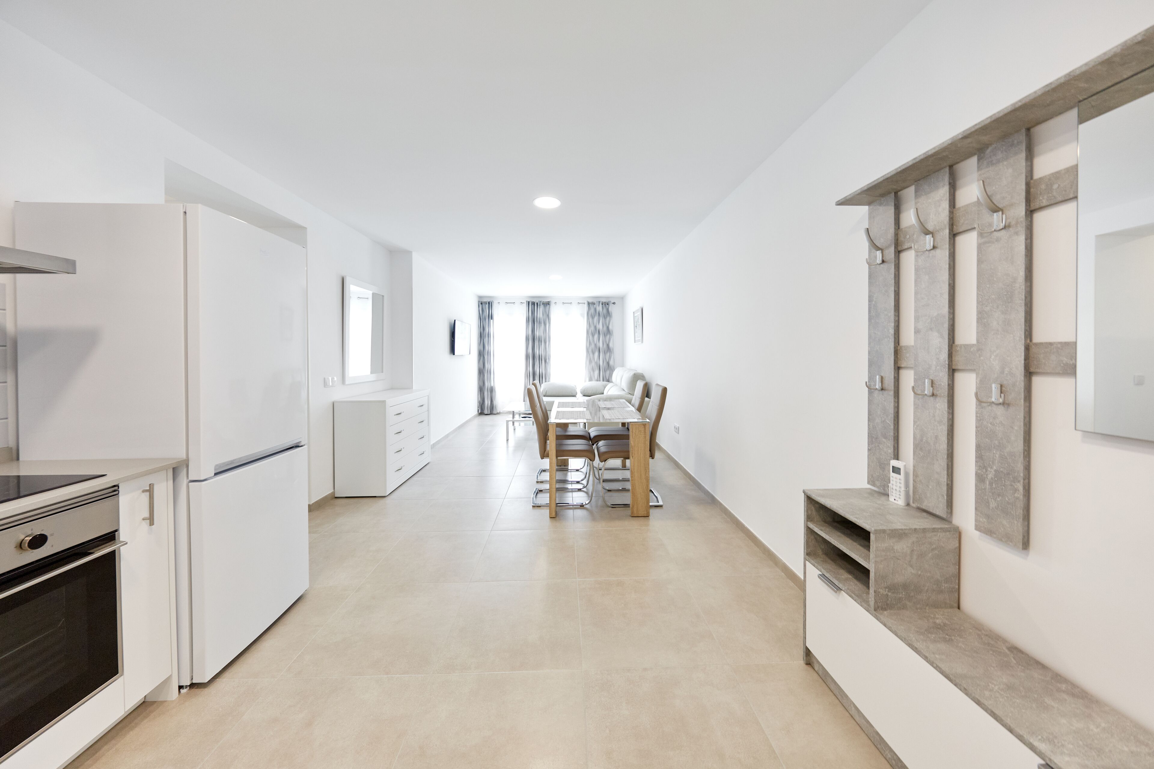 A modern kitchen with white appliances, a dining area with a table and chairs, and a hallway leading to a bedroom.