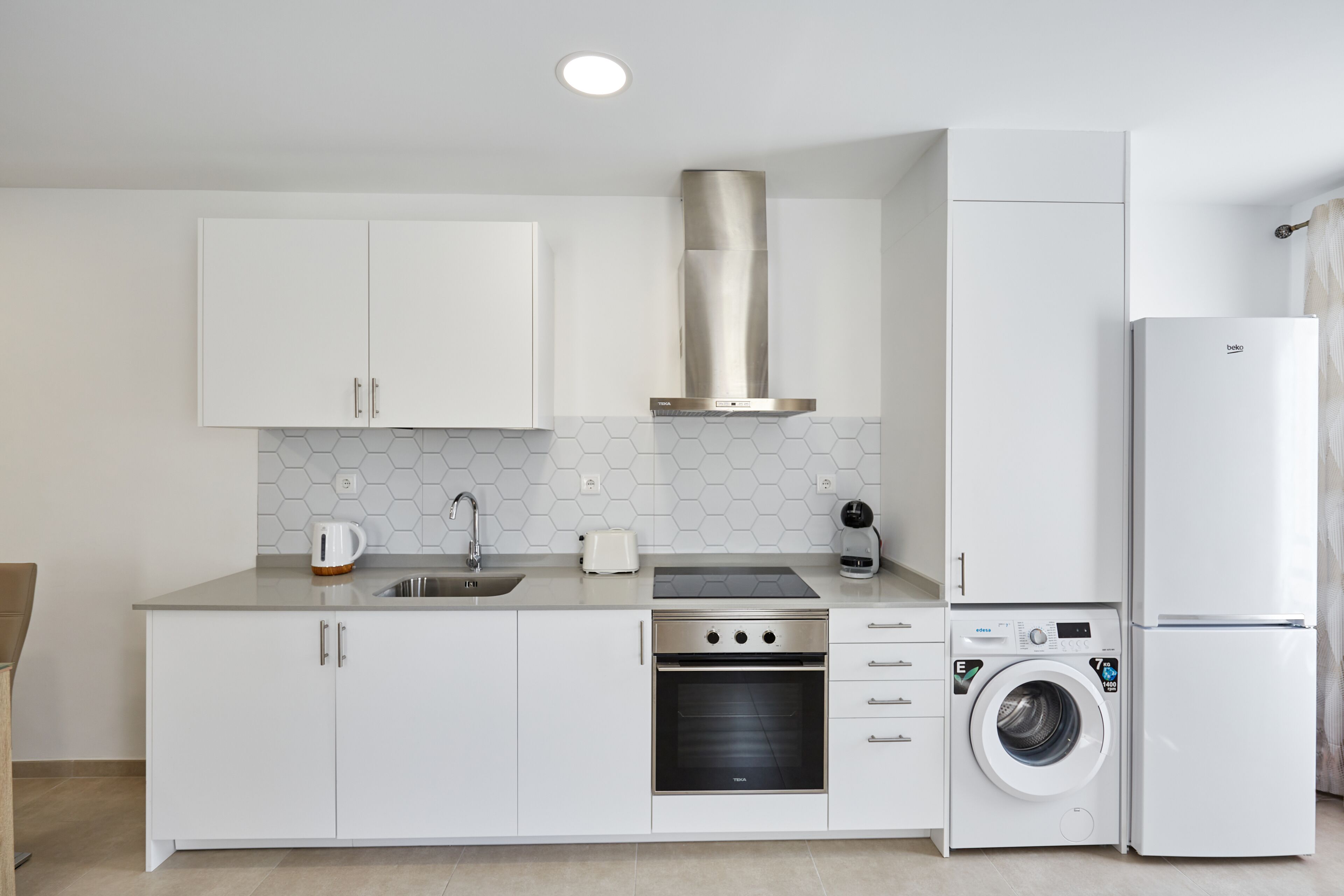 Una cocina compacta con armarios blancos, una estufa de acero inoxidable, horno empotrado, lavadora y refrigerador.
