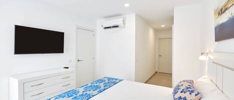 A bedroom with a bed, a TV mounted on the wall, a white dresser, and an air conditioning unit.