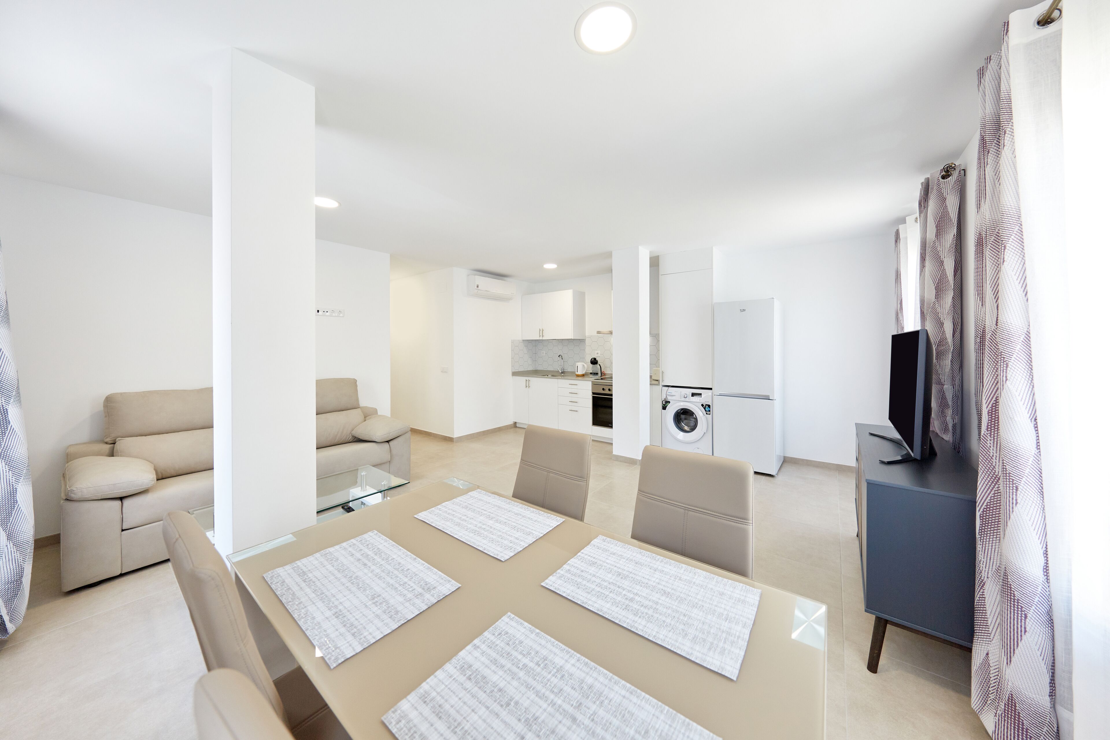 Una sala de estar moderna con un sofá, una mesa de comedor con manteles individuales, un televisor sobre un mueble bajo y una cocina al fondo.