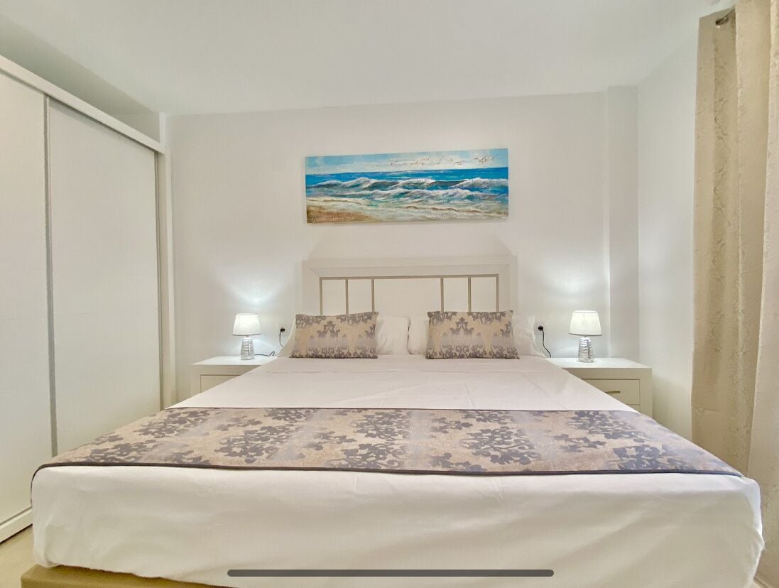 Uma cama bem arrumada com um edredom estampado, duas luminárias de cabeceira e uma pintura de uma paisagem praiana acima da cama.