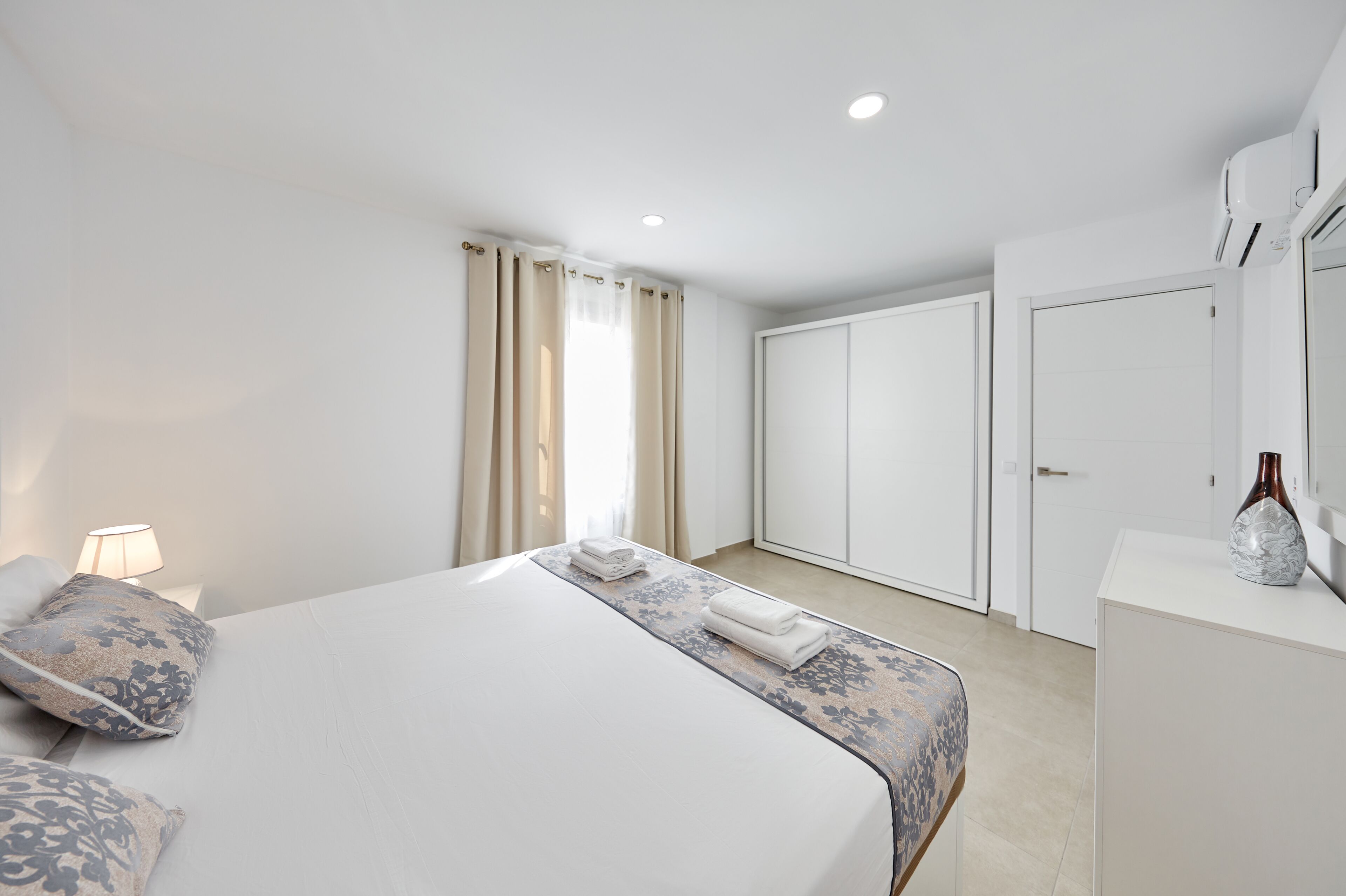 A modern hotel room with a large bed, a bedside table with a lamp, a wardrobe, and a mirror.