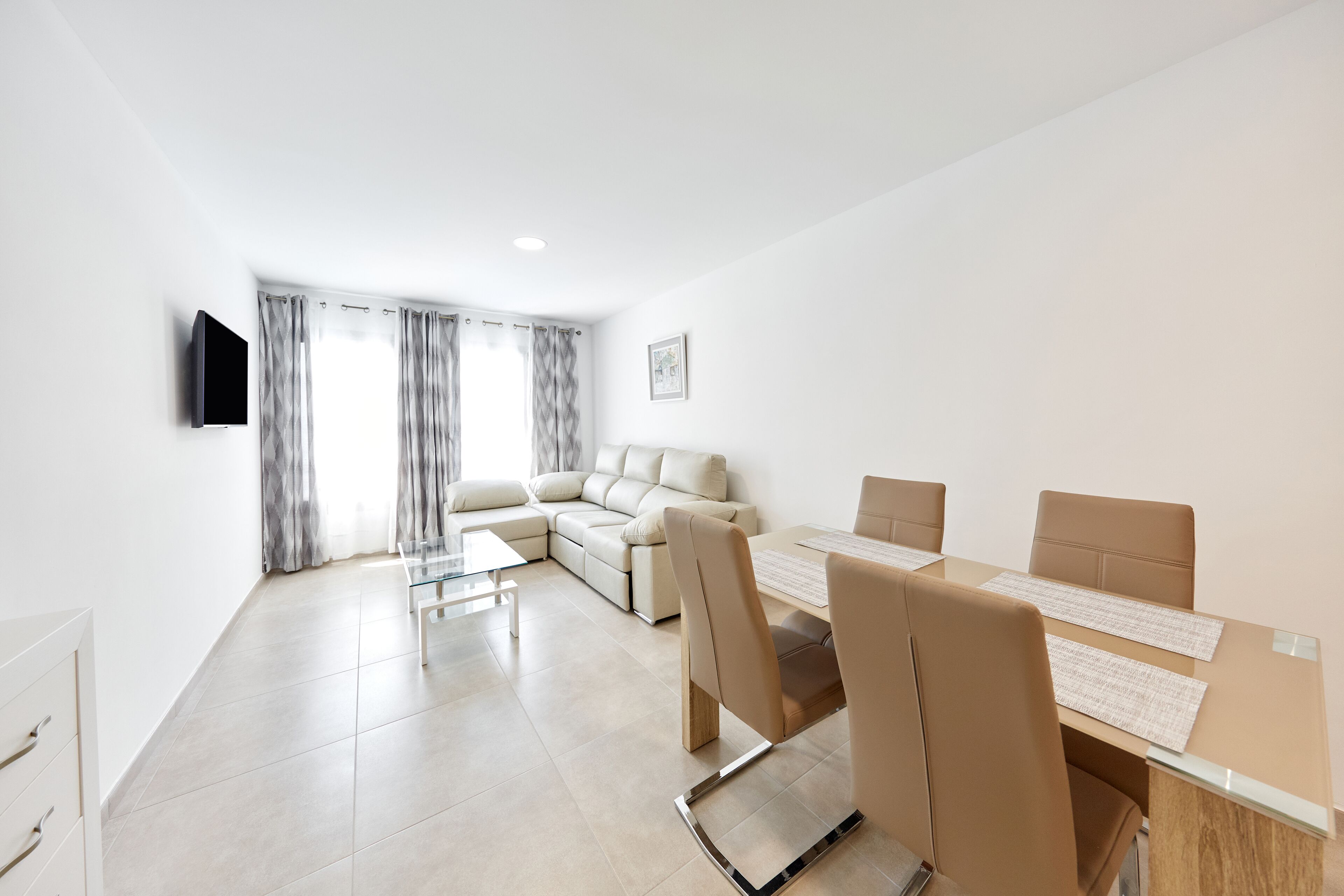 A modern living room with a sofa, a dining table with chairs, a flat-screen TV mounted on the wall, and a window with curtains.