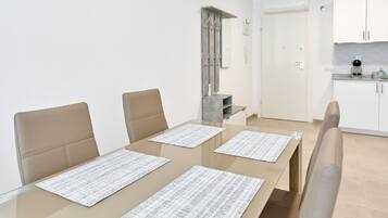A modern dining area with a glass table, beige chairs, and a kitchen in the background.
