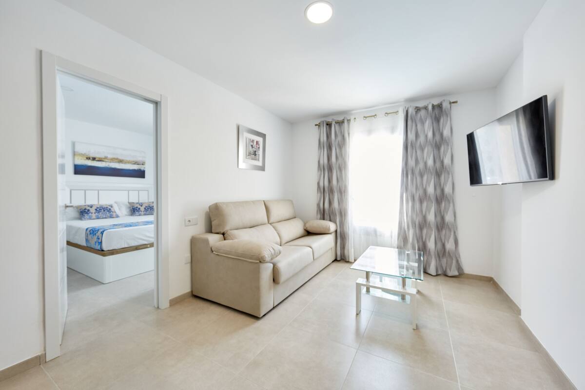 Een moderne woonkamer met een beige bank, een glazen salontafel en een flatscreen televisie aan de muur.