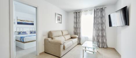 A modern living room with a beige sofa, a glass coffee table, and a flat-screen TV mounted on the wall.