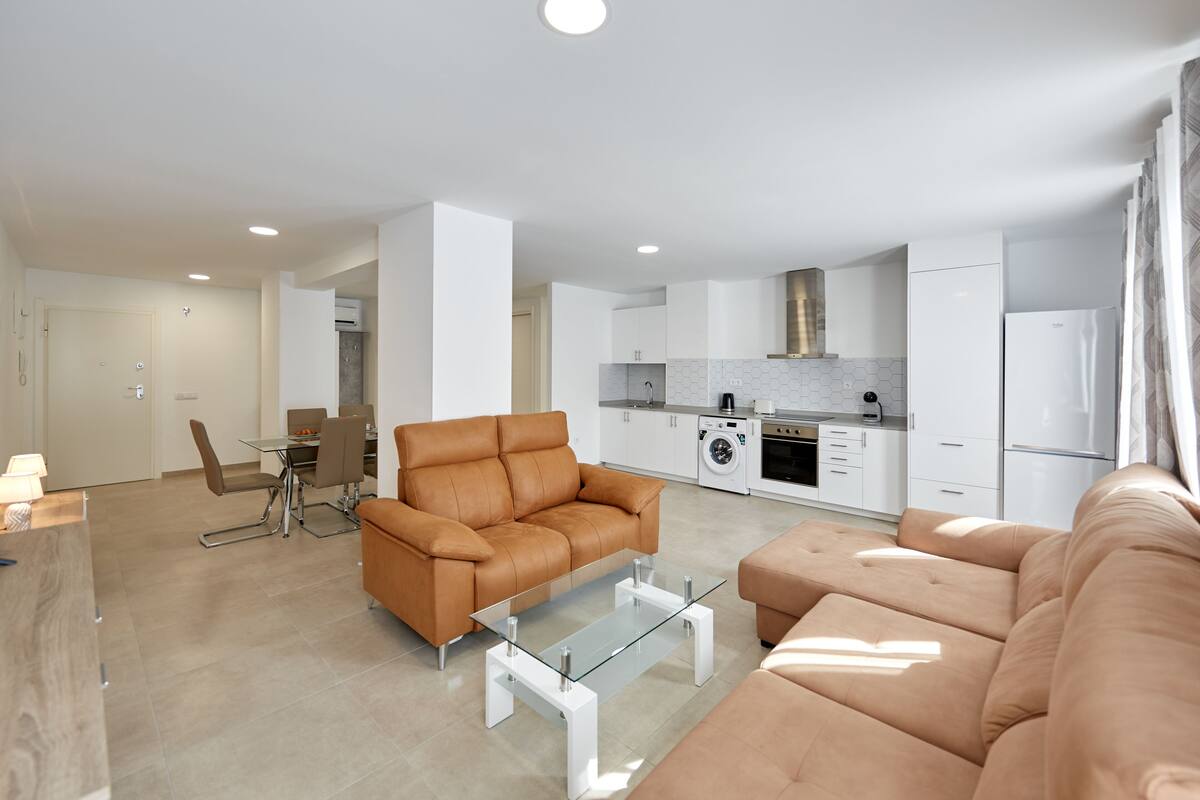 Een moderne woonkamer met een bruine leren bank, een glazen salontafel en een eethoek met een tafel en stoelen.