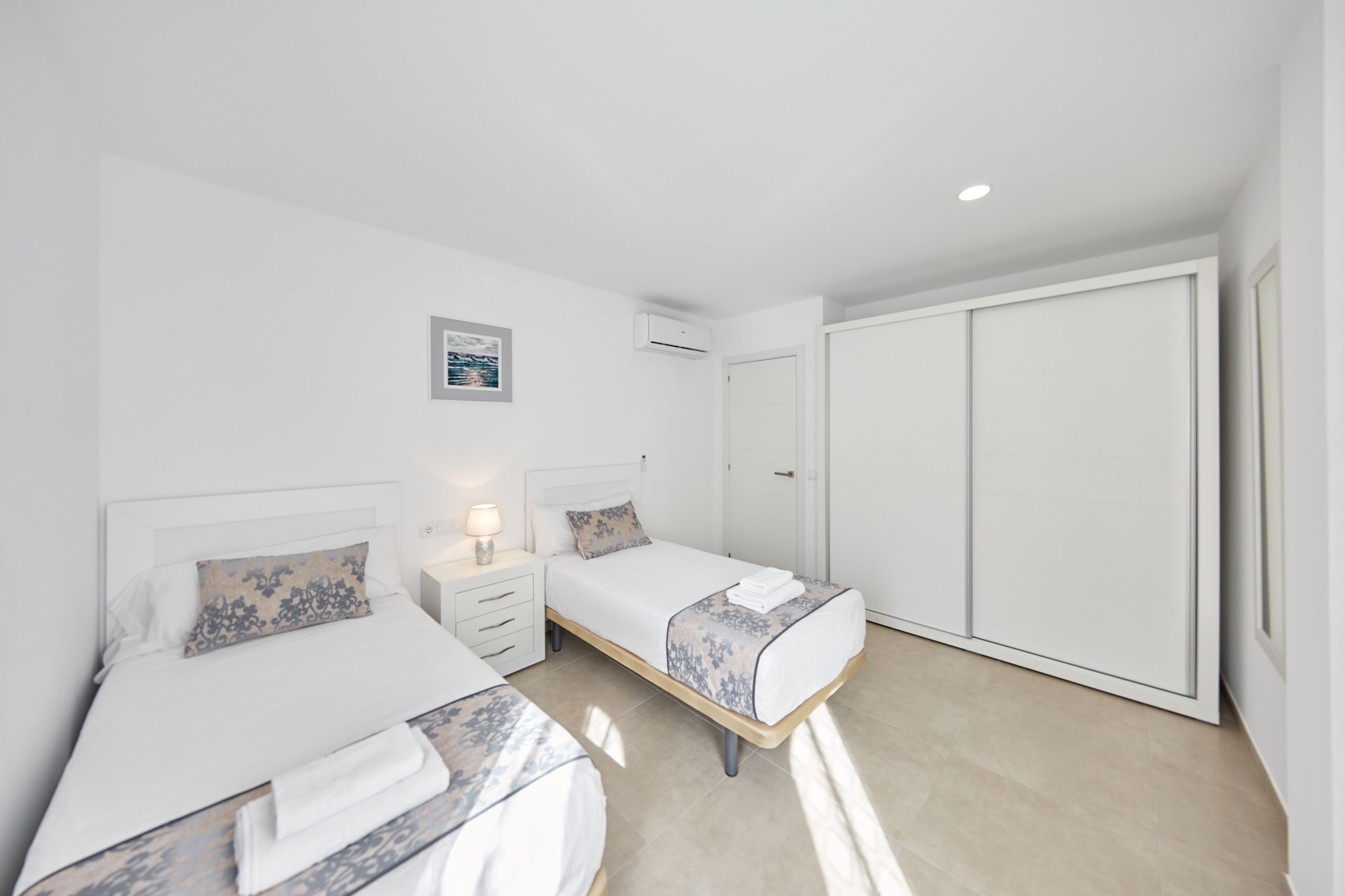 Habitación de hotel con dos camas individuales, una mesita de noche con lámpara, un armario y un equipo de aire acondicionado.