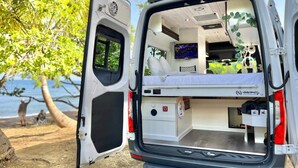 Een witte bus met open achterkant, waar een bed, een kleine keuken en een televisie aan de wand te zien zijn.
