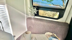 Een gezellig interieur met een zachte zitting, een raam met uitzicht op het strand, een klein tafeltje met een boek en een mok, en een decoratieve plant.