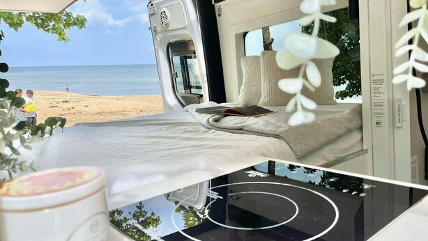 Een camper met een bed, een raam en uitzicht op het strand.