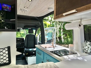 A compact RV interior with a kitchenette, a seating area, and a TV displaying 'The Roku Channel'.