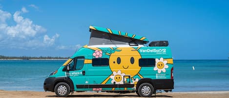 Ein türkisfarbener Campervan mit Sonn- und Blumenmotiv parkt am Strand.