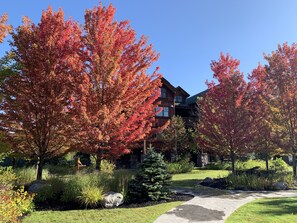 A lodge with a stone facade, a wooden deck, and a well-maintained garden with autumn foliage. - 15-45% Off- Whiteface Lodge- Pools, Hot tubs, Spa, Game Room & Theatre! (Lake Placid)