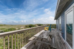 A wooden deck with a table and chairs, overlooking a beach with dunes and a lighthouse in the distance.