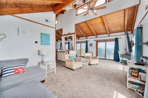 A spacious living area with a high ceiling, wooden beams, a sofa with a striped pillow, a small table, and a bookshelf.