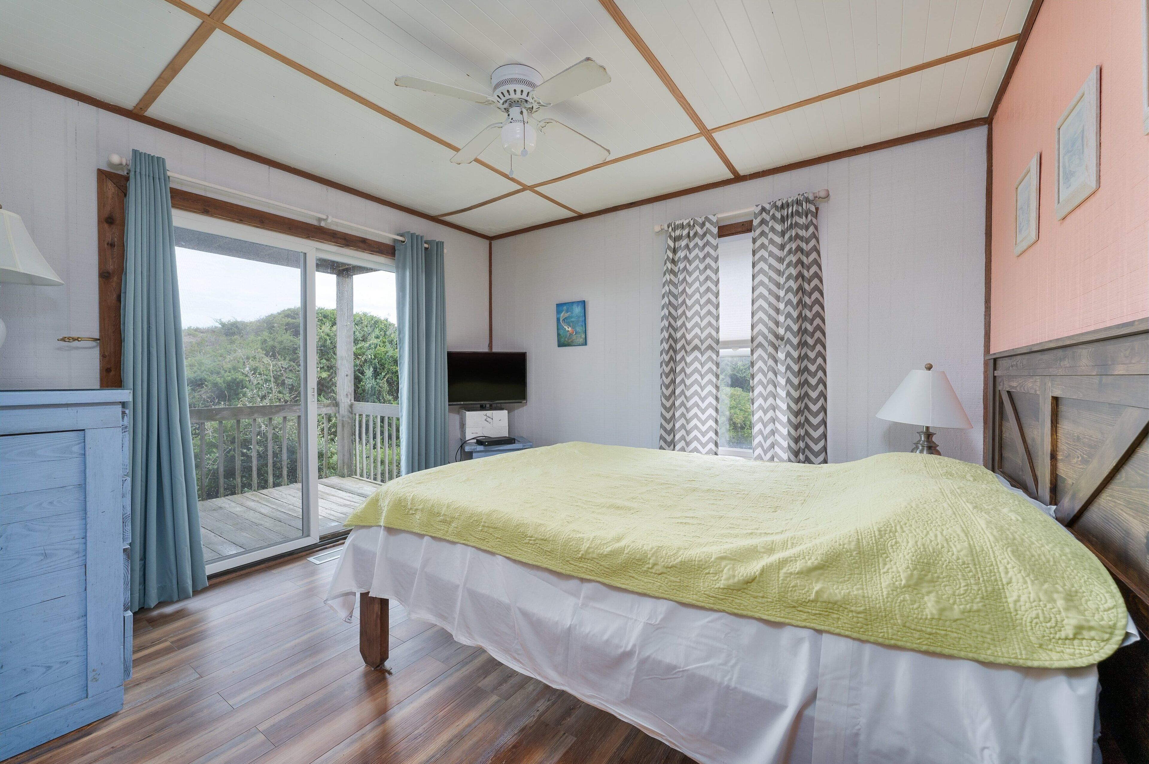 A bedroom with a bed, a television, a ceiling fan, a window with curtains, and a balcony.