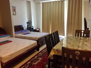 A hotel room with two beds, a dining table, chairs, a fan, and a window with curtains.
