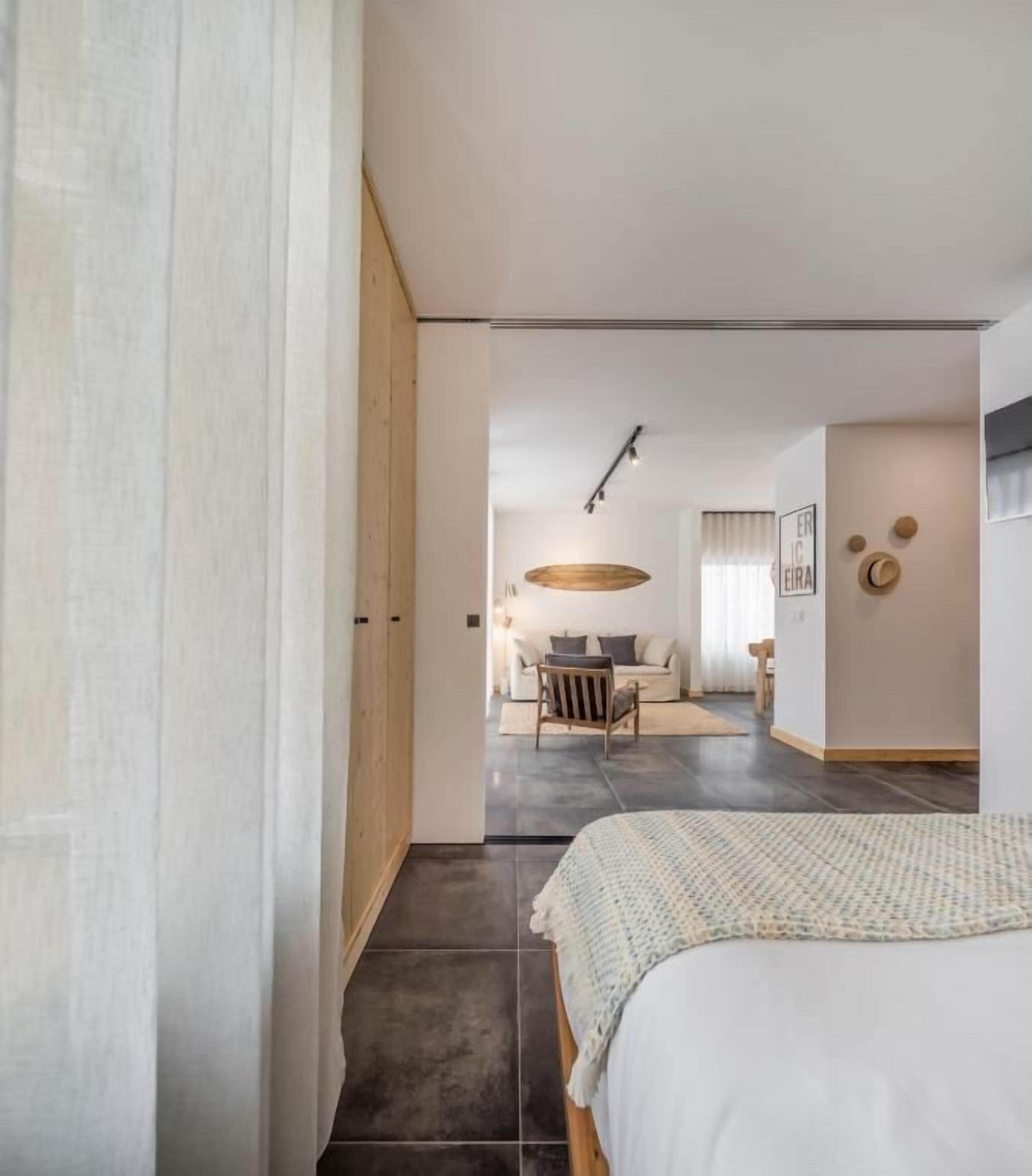 A modern hotel room with a large bed, a wooden wardrobe, a dining area with a table and chairs, and a kitchen in the background.