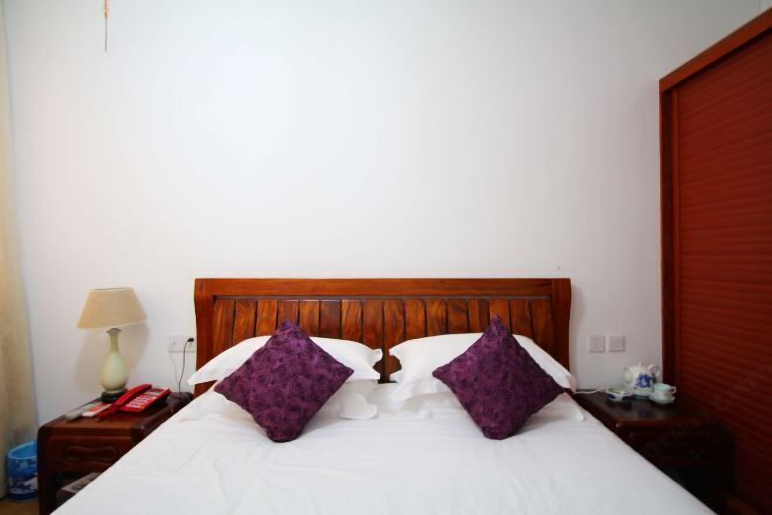 A hotel room with a wooden headboard, a bed with white linens and purple pillows, a bedside table with a lamp, a red telephone, and a small table with a cup and saucer.