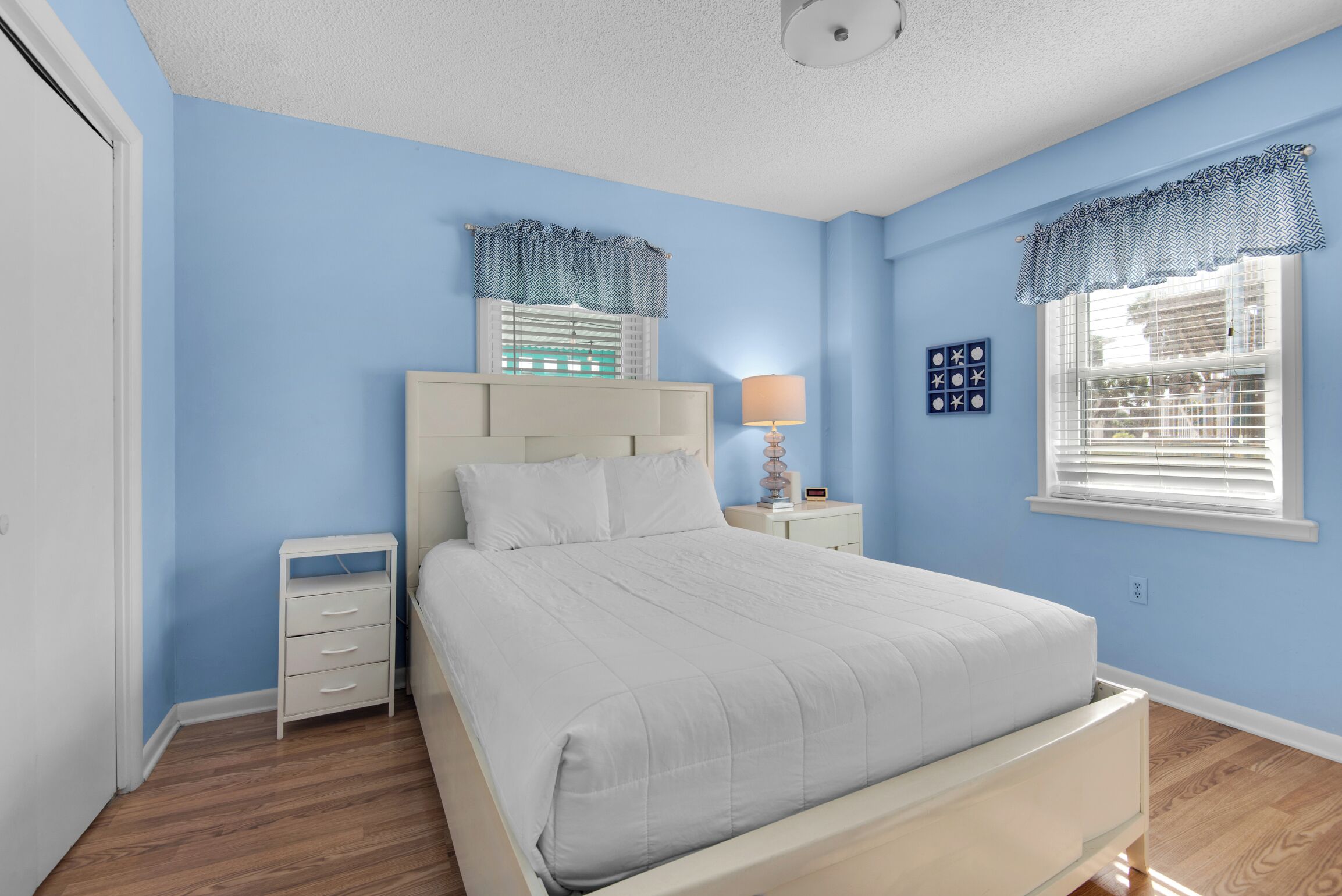 Une chambre à coucher comprenant un lit, une table de chevet, une commode et une fenêtre avec des stores.
