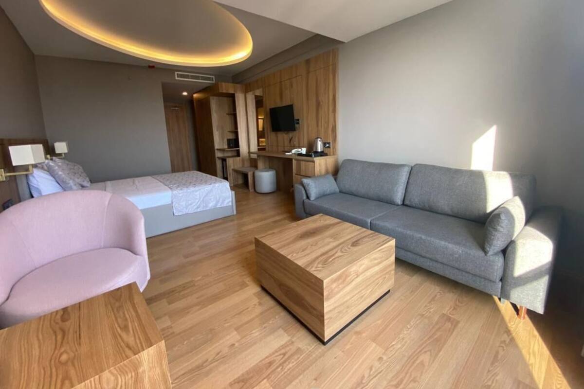 Una habitación de hotel moderna con piso de madera, un sofá gris, un sillón rosa, una mesa de centro de madera, una cama con ropa blanca, un televisor montado en la pared y un mueble de madera.