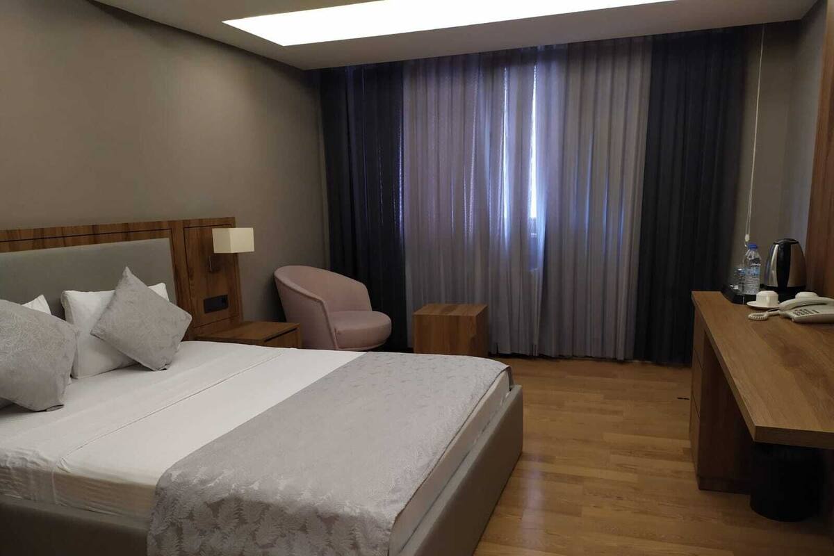 Habitación de hotel con una cama grande, un escritorio, una silla, una mesita de noche y un ventanal con cortinas.