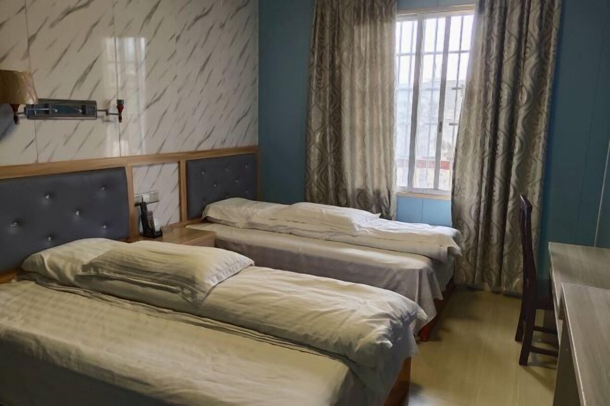 Habitación de hotel con dos camas, una ventana con cortinas, cabecero de madera y una mesita pequeña.