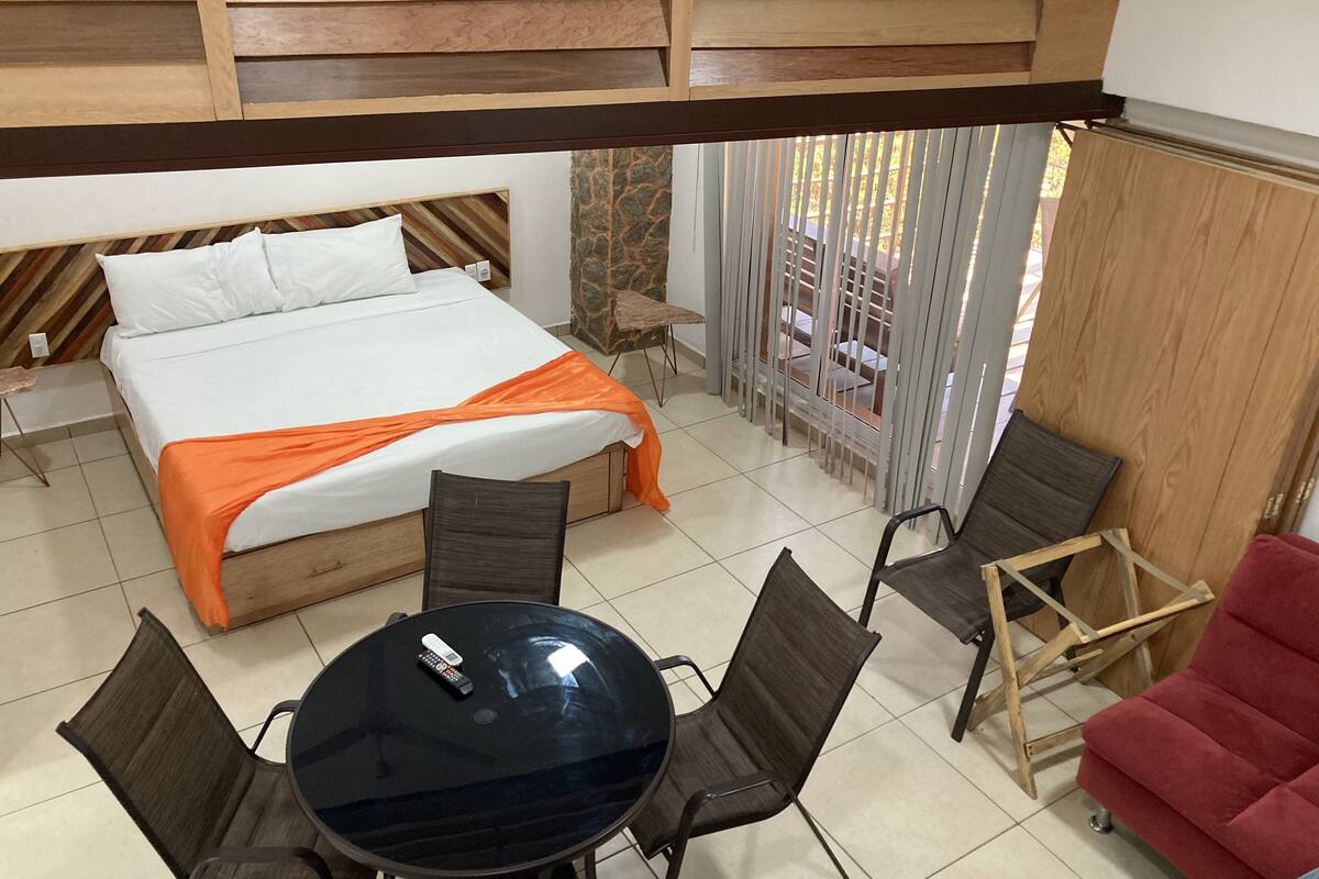 Quarto de hotel com uma cama, uma mesa redonda com cadeiras, uma poltrona vermelha e uma varanda com vista.