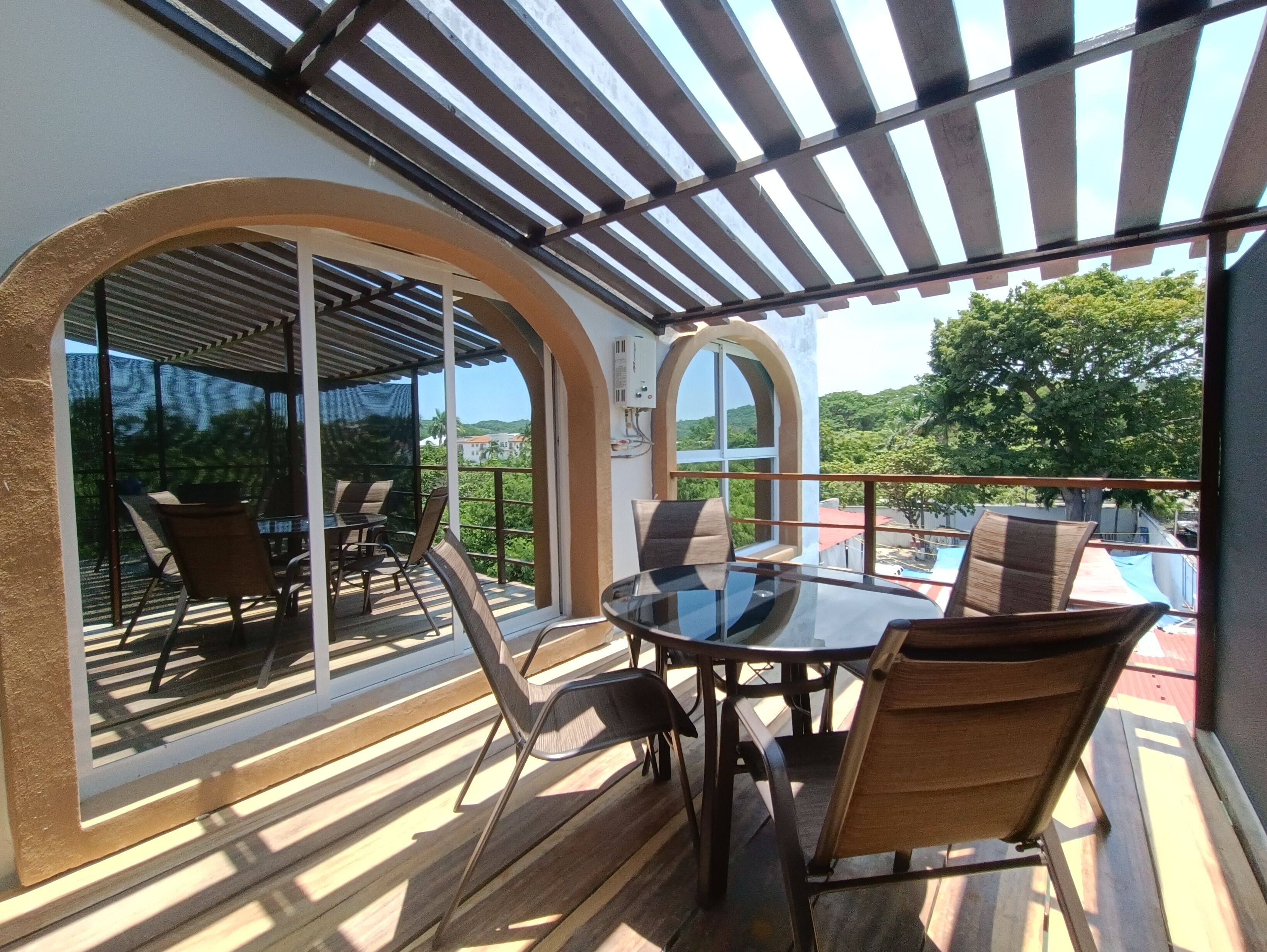 An outdoor patio with a glass door leading to a balcony, furnished with a table and chairs, and a pergola overhead.