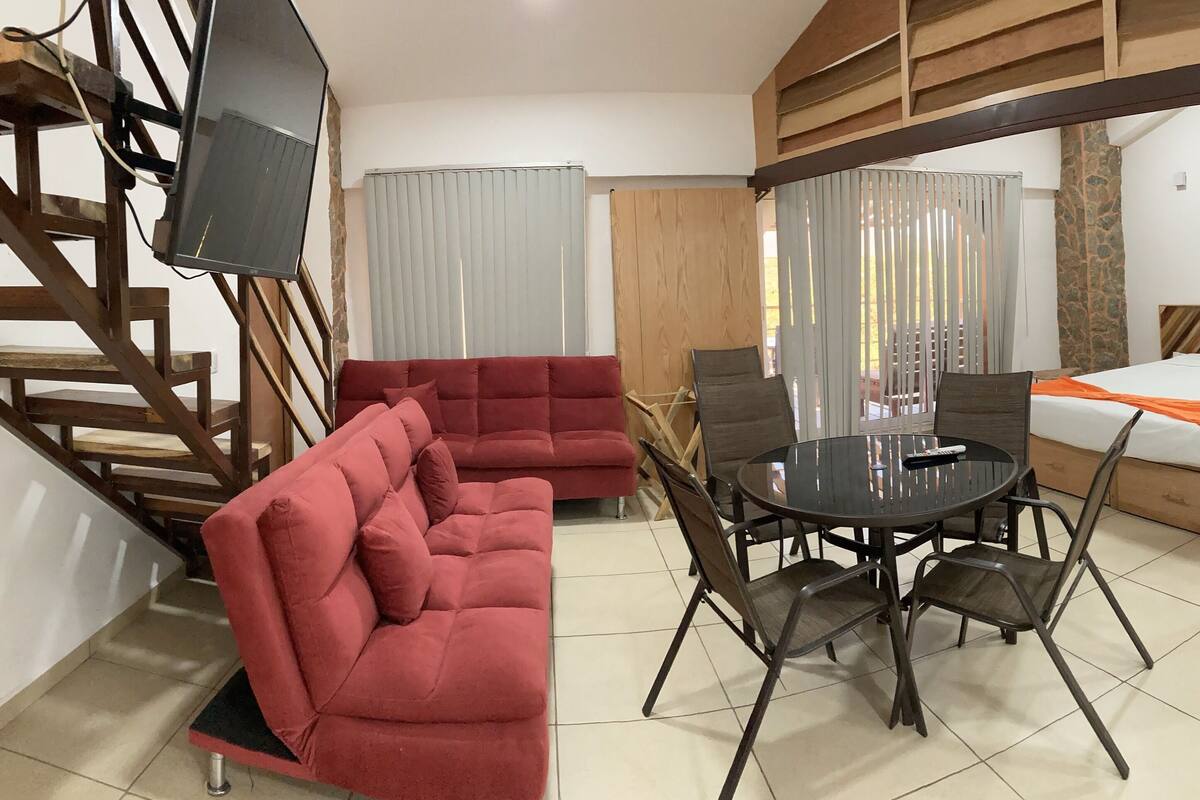 Quarto de hotel com sofá, mesa de jantar com cadeiras, cama e televisão fixada numa estrutura de madeira.