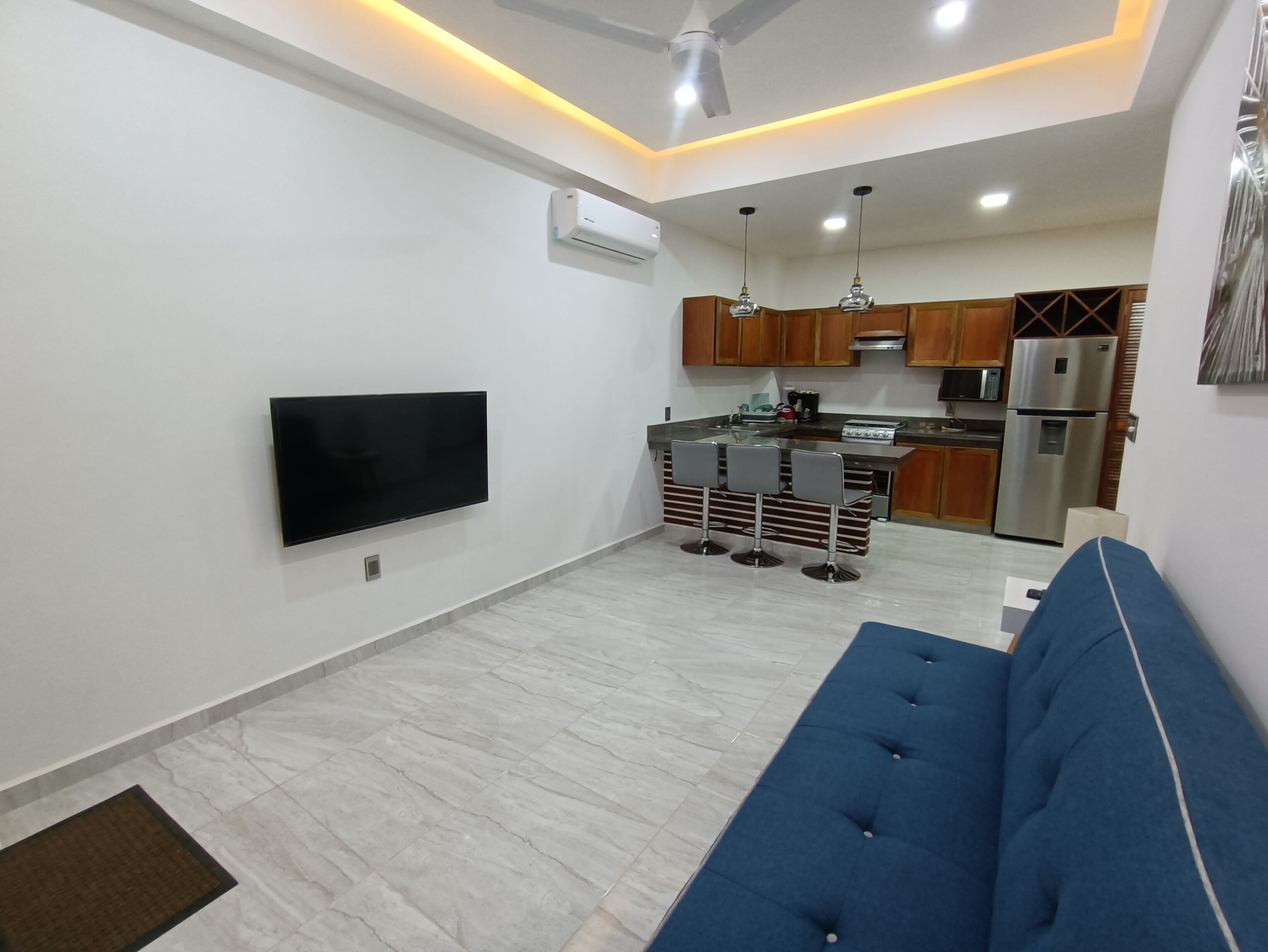 Una sala de estar moderna con un televisor de pantalla plana, un sofá azul y una cocina con armarios de madera y un refrigerador de acero inoxidable.