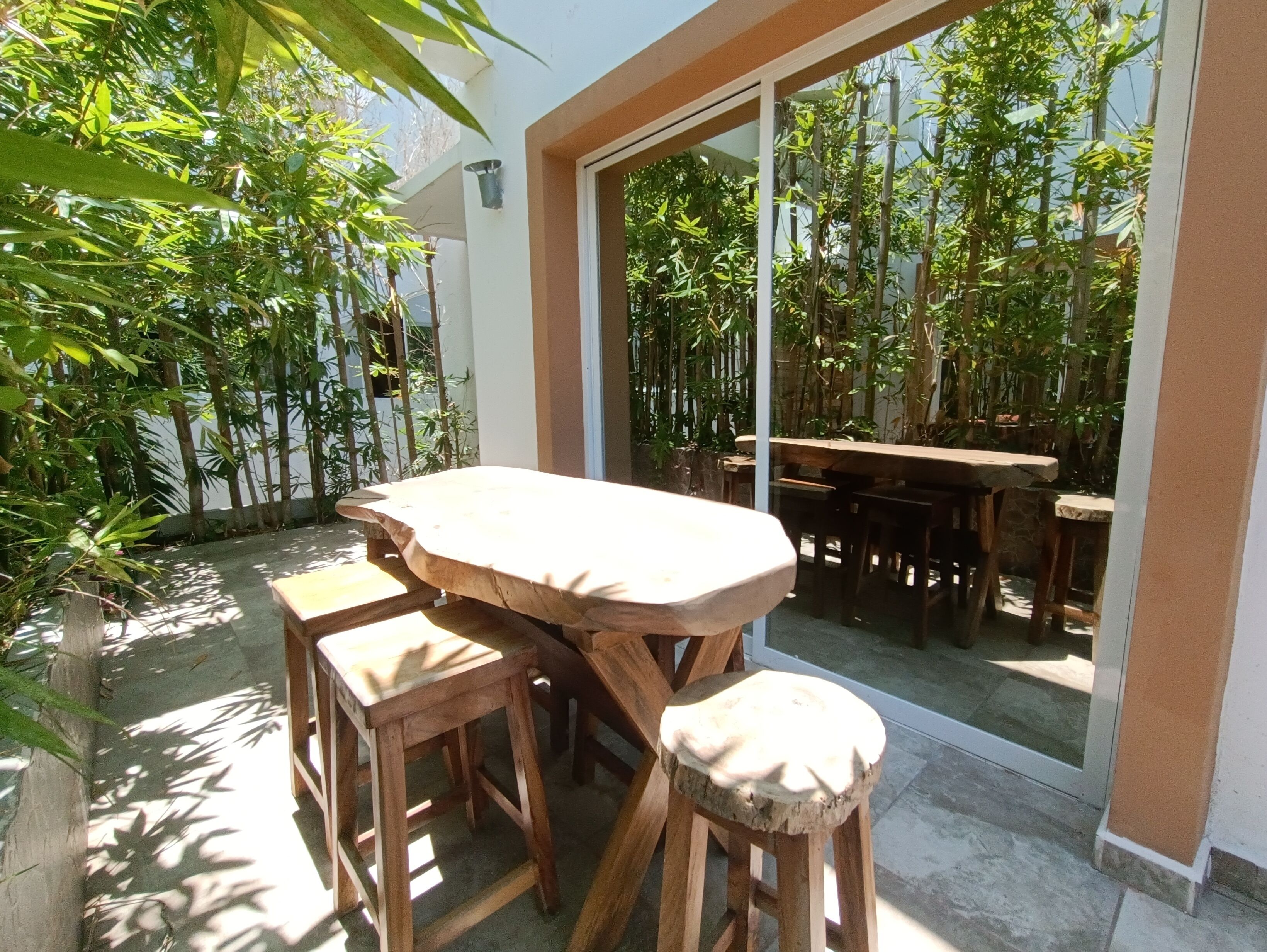 A wooden table and stools in a sunlit outdoor area with bamboo plants and a glass door leading to a shaded patio.