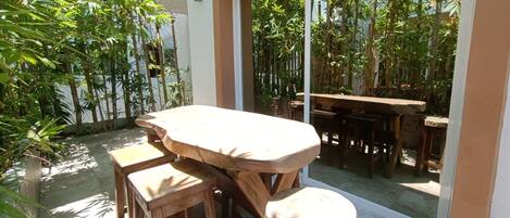 A wooden table and stools in a sunlit outdoor area with bamboo plants and a glass door leading to a shaded patio.