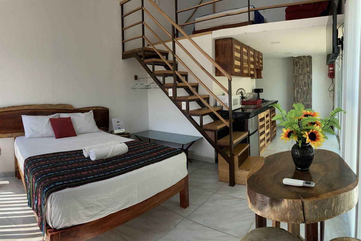 Quarto com cama de madeira, uma pequena mesa de estudo, escada de madeira e área de cozinha.