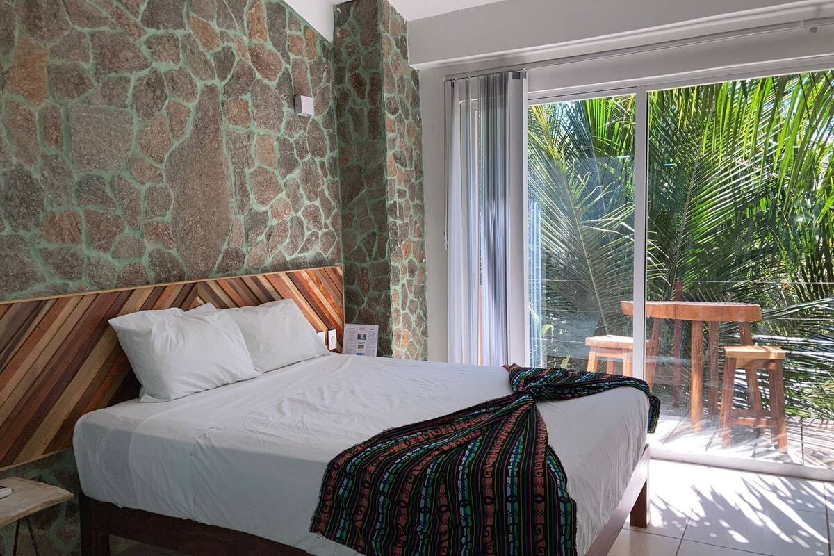 Quarto com parede de pedra, cabeceira de madeira, cama com roupa de cama branca e manta colorida, vista para palmeiras por uma porta de vidro deslizante.